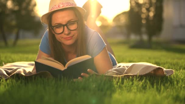 Девушка в очках читает книгу, лежащую на одеяле в парке на закате — стоковое видео