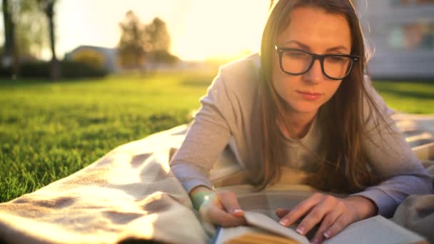 Menina de óculos lendo livro deitado em um cobertor no parque ao pôr do sol — Vídeo de Stock