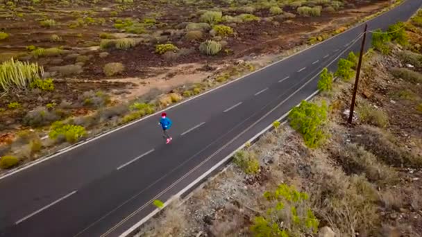 Vista aérea de la mujer corre a lo largo de la carretera de asfalto desierta al atardecer. Montañas en el fondo — Vídeos de Stock