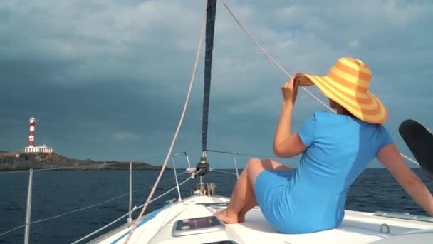 Vrouw in een gele hoed en een blauwe jurk meisje rust aan boord van een jacht in de buurt van de vuurtoren op het zomerseizoen op oceaan — Stockvideo