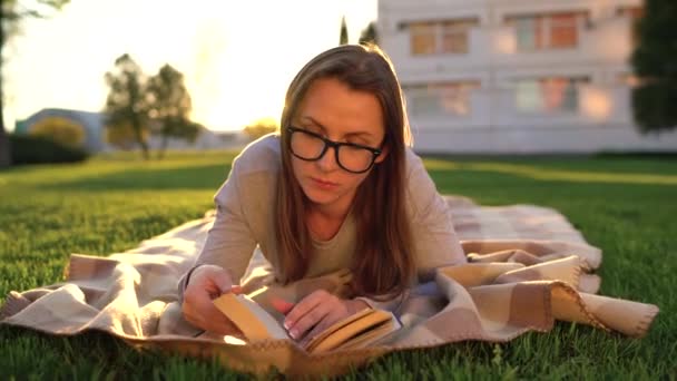 Девушка в очках читает книгу, лежащую на одеяле в парке на закате — стоковое видео