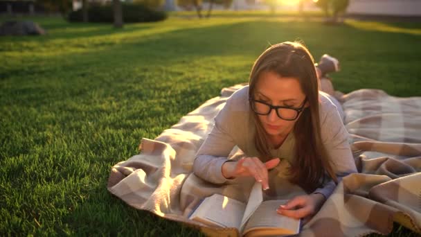Девушка в очках читает книгу, лежащую на одеяле в парке на закате — стоковое видео