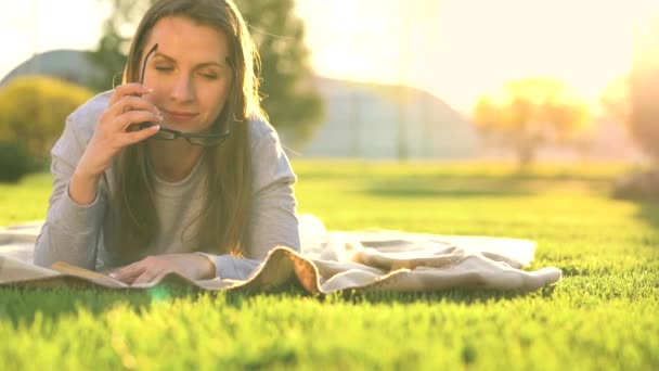 Szemüveg-olvasó könyv fekve egy takarót a parkban a naplemente — Stock videók