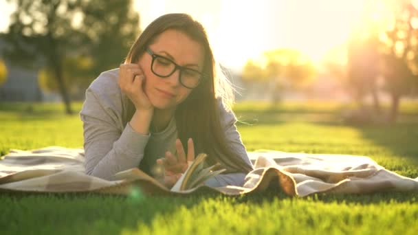 Девушка в очках читает книгу, лежащую на одеяле в парке на закате — стоковое видео