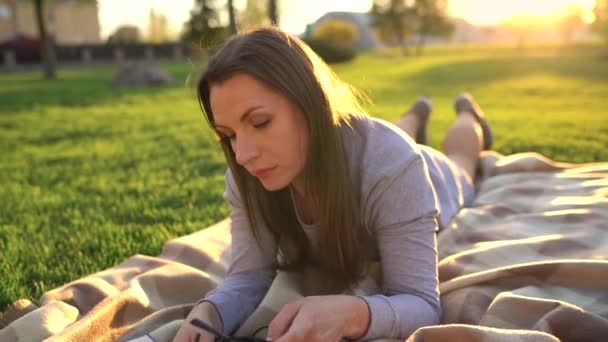 Menina de óculos lendo livro deitado em um cobertor no parque ao pôr do sol — Vídeo de Stock