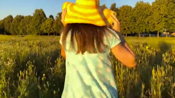 Belleza chica corriendo en un sombrero amarillo en el campo de trigo verde sobre el cielo puesta del sol. Concepto de libertad. Campo de trigo al atardecer — Vídeos de Stock