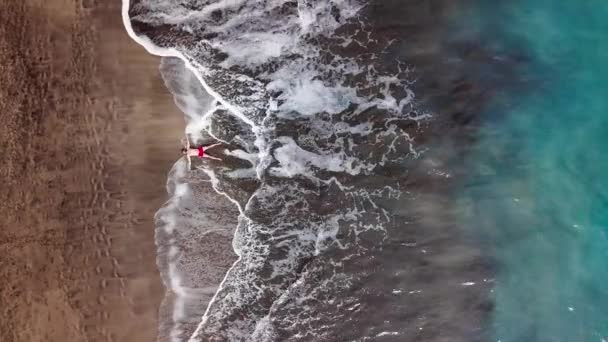 Vista aérea de un hombre en pantalones cortos rojos se encuentra en la playa volcánica negra desierta en una pose estelar. Imágenes aéreas de drones de olas marinas que llegan a la orilla — Vídeos de Stock