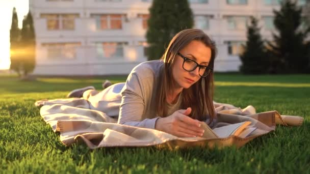 Dívka v brýlích čtení knihy vleže na dece v parku při západu slunce — Stock video