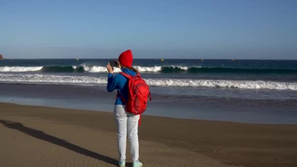 Активні мандрівного жінка прогулянки по пляжу і робить фото її смартфон. Кавказький молода жінка з рюкзака на Тенеріфе, Канарські острови, Іспанія — стокове відео