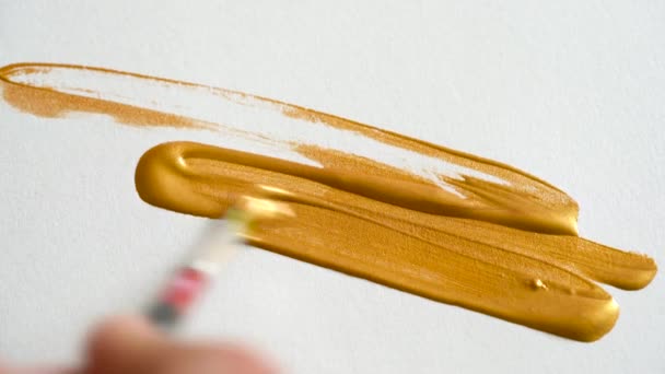 Dibujo con pincel sobre papel blanco con pintura acrílica dorada brillante de cerca — Vídeos de Stock