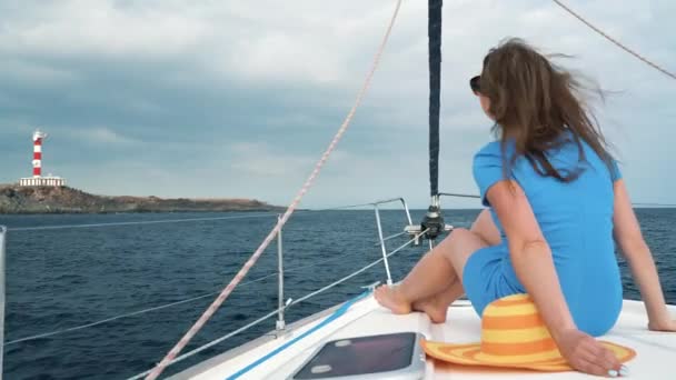 Mujer con un sombrero amarillo y vestido azul descansa a bordo de un yate cerca del faro en la temporada de verano en el océano — Vídeo de stock