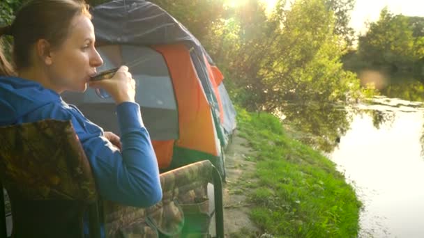 Woman is drinking tea in an armchair near a tent on the river bank — Stock Video