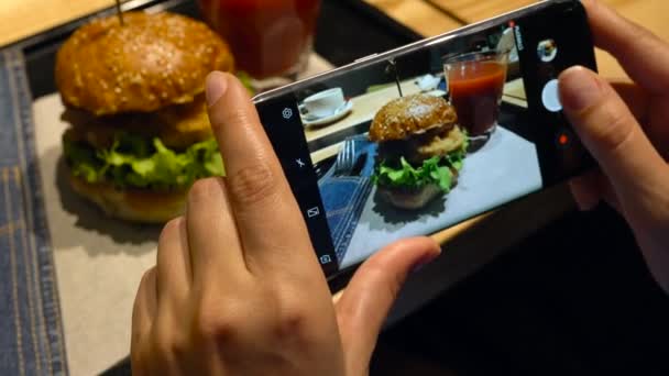 Κορίτσι κάνει μια φωτογραφία του burger και χυμό ντομάτας σε ένα smartphone σε ένα καφέ εσωτερικη — Αρχείο Βίντεο