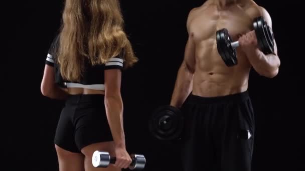 Atlético hombre y mujer flexiona sus manos con mancuernas, entrenando sus bíceps sobre un fondo negro en el estudio — Vídeo de stock