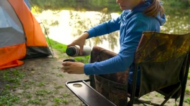 Kadın bir koltuk nehir kıyısında bir çadırın yakınında çay içiyor