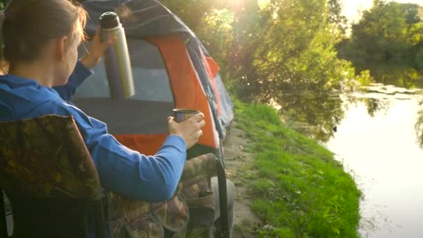 Woman is drinking tea in an armchair near a tent on the river bank — Stock Video