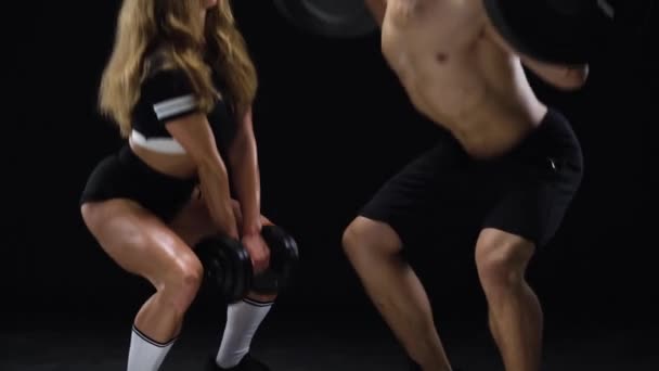 Athletic man and woman crouch with extra weight, training their legs and buttocks on a black background in studio — Stock Video