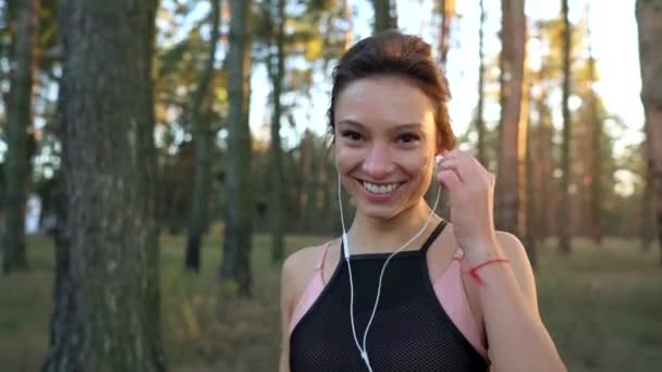 Nahaufnahme einer lächelnden Frau, die Kopfhörer in die Ohren steckt und bei Sonnenuntergang durch einen herbstlichen Wald läuft — Stockvideo