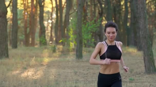 Primer plano de mujer con auriculares corriendo a través de un bosque otoñal al atardecer. Movimiento lento — Vídeos de Stock