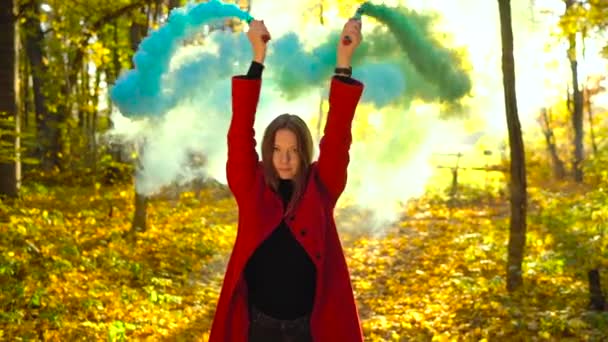Hermosa chica con un abrigo rojo sostiene humo de color en sus manos y camina a través del bosque amarillo de otoño. Movimiento lento — Vídeo de stock