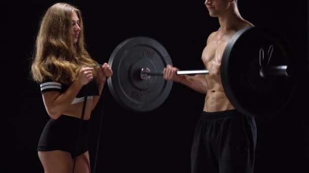 Homem e mulher atléticos flexiona as mãos com halteres e com um sino, treinando seus bíceps em um fundo preto no estúdio — Vídeo de Stock