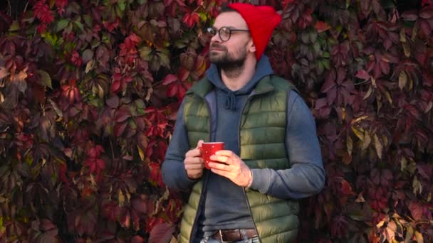 El hombre con una gorra roja con una taza en las manos está bebiendo té o café al aire libre y usando un teléfono inteligente en un cálido día de otoño. Pausa para café . — Vídeos de Stock