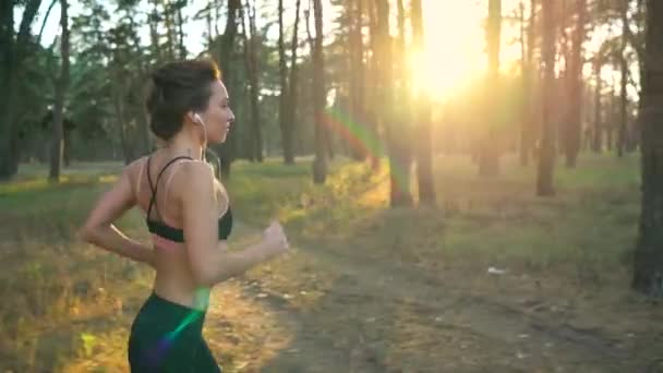 Primer plano de mujer con auriculares corriendo a través de un bosque otoñal al atardecer. Movimiento lento — Vídeos de Stock
