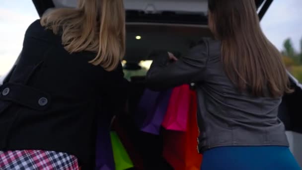 Två glada kvinnor efter shopping sätta väskor i bakluckan på bilen, sitta i den och gör dig redo att köra iväg — Stockvideo