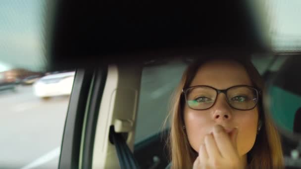 Mooi meisje in in glazen schildert lippen in de auto en en kijkt in de achteruitkijkspiegel — Stockvideo