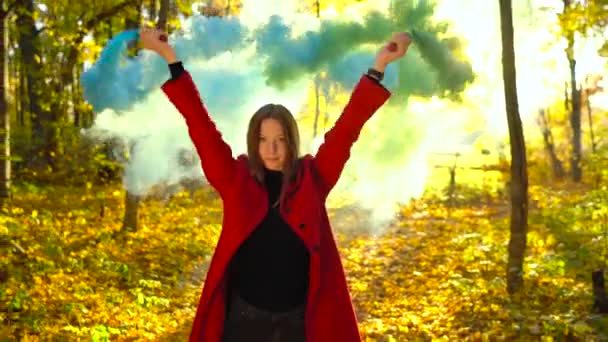 Hermosa chica con un abrigo rojo sostiene humo de color en sus manos y camina a través del bosque amarillo de otoño — Vídeos de Stock