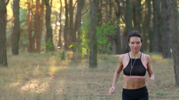 Primer plano de la mujer con auriculares corriendo a través de un bosque de otoño al atardecer — Vídeos de Stock