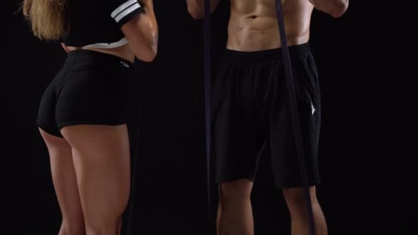Atletický muž a žena, která dělá cvičení na biceps s elastickým pásem na černém pozadí v studio — Stock video