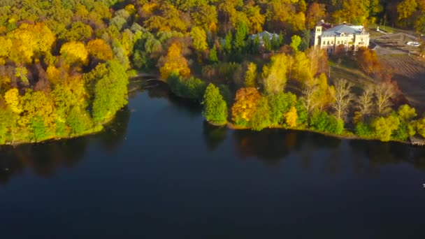 Вид с воздуха на озеро, усадьбу и яркий осенний лес на его берегу. Лес отражается на поверхности озера — стоковое видео