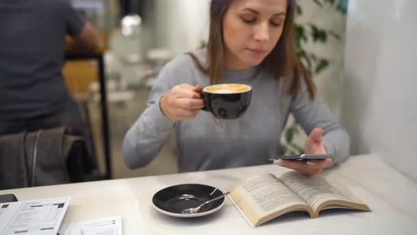 Красивая женщина проводит время в кафе: читает книгу, разговаривает по телефону и пьет кофе — стоковое видео