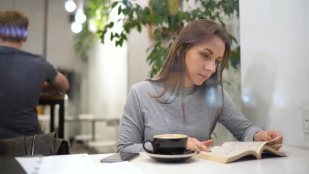 Schöne Frau verbringt ihre Zeit in einem Café: ein Buch lesen und Kaffee trinken — Stockvideo