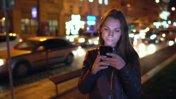 Mujer atractiva utiliza un teléfono inteligente mientras camina por las calles de la ciudad de la noche — Vídeos de Stock