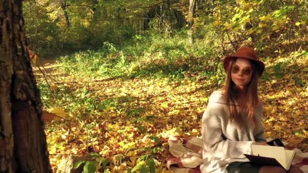 Hermosa mujer se sienta en un cuadros en el bosque de otoño, toma notas en un cuaderno y disfruta del buen tiempo. Movimiento lento — Vídeo de stock