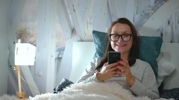 Mujer en gafas se encuentra en una cama y utiliza un teléfono inteligente antes de acostarse — Vídeos de Stock