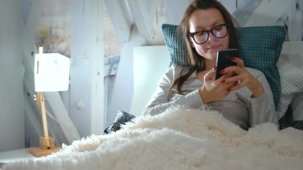 Γυναίκα γυαλιά βρίσκεται σε ένα κρεβάτι και χρησιμοποιεί ένα smartphone πριν τον ύπνο — Αρχείο Βίντεο