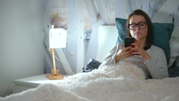 Mulher de óculos fica em uma cama e usa um smartphone antes de dormir — Vídeo de Stock