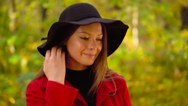 Retrato de una hermosa chica sonriente en un sombrero negro con una hoja de arce amarillo en primer plano en el bosque de otoño — Vídeos de Stock