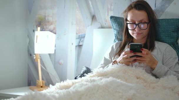 Frau mit Brille liegt im Bett und nutzt Smartphone vor dem Schlafengehen — Stockvideo