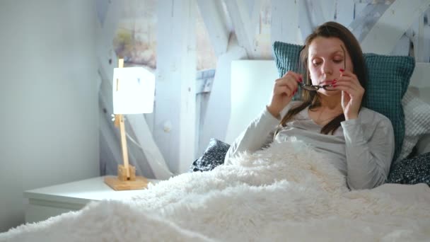 Mujer en gafas se encuentra en una cama y utiliza un teléfono inteligente antes de acostarse — Vídeo de stock