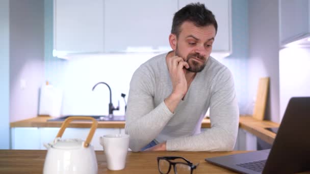 Бородатый сонный мужчина завтракает на кухне и пользуется ноутбуком — стоковое видео