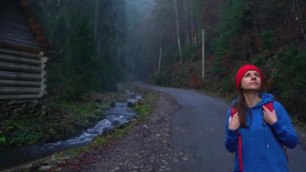 Femme voyageur avec un sac à dos marche sur la route dans les montagnes le long de la rivière et admire le paysage environnant — Video