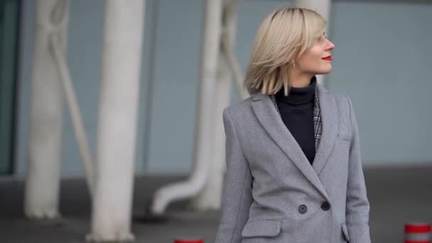 Blonde fille dans un manteau gris va sur le fond d'un bâtiment en verre moderne — Video