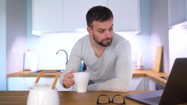 Homem com sono barbudo tomando café da manhã na cozinha e usando um laptop — Vídeo de Stock