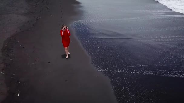 Vue aérienne d'une fille en robe rouge marchant sur la plage avec du sable noir. Tenerife, Îles Canaries, Espagne — Video