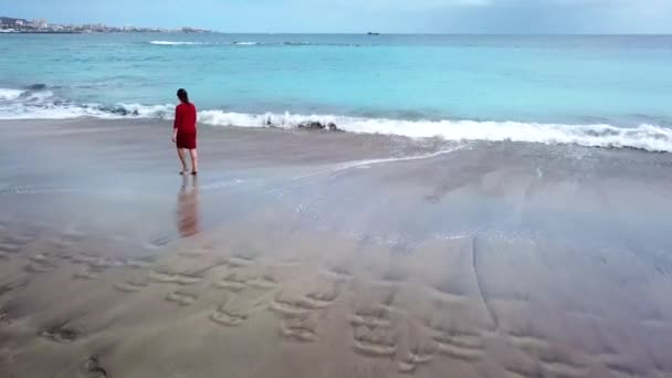 Flygfoto över en flicka i en röd klänning promenader på stranden med svart sand. Teneriffa, Kanarieöarna, Spanien — Stockvideo