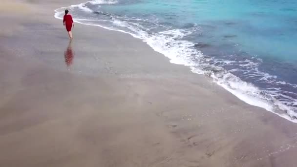 Vista aérea de uma menina em um vestido vermelho andando na praia com areia preta. Tenerife, Ilhas Canárias, Espanha — Vídeo de Stock